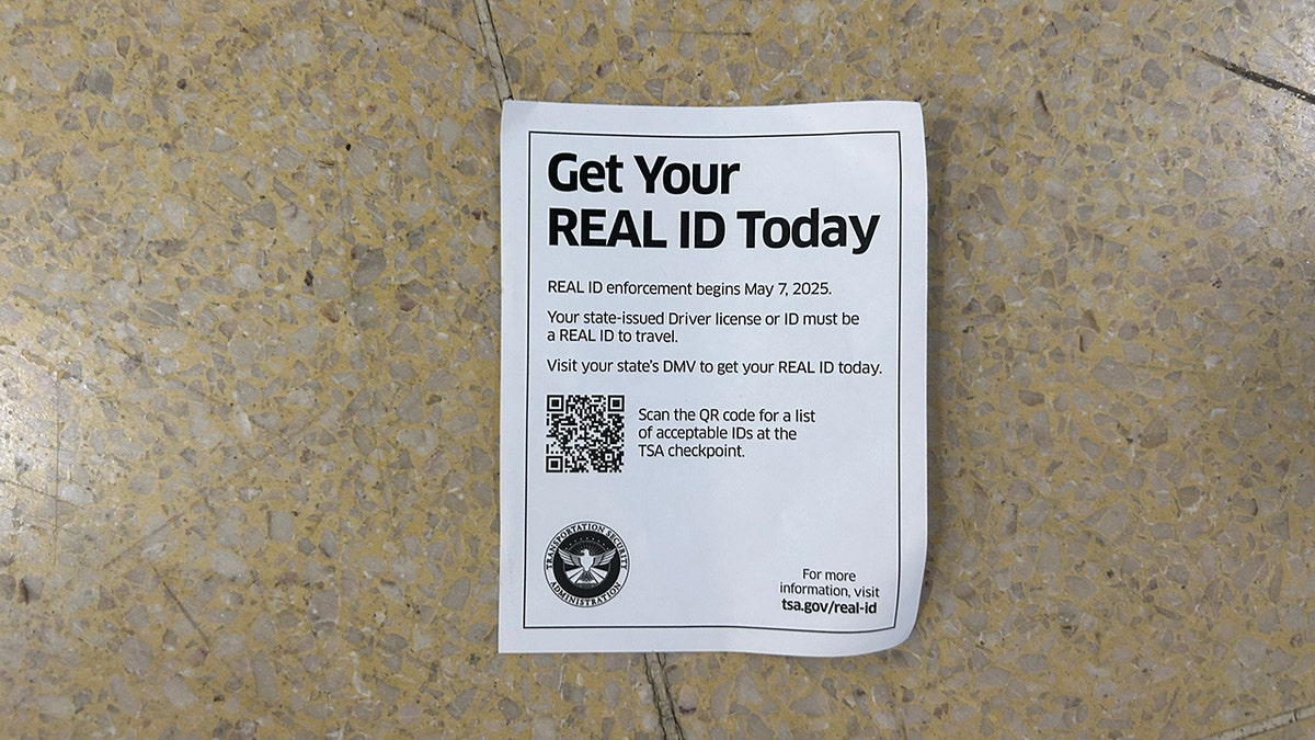 Paper flyer on the ground encouraging travelers to upgrade to a REAL ID. It reads: "Get Your REAL ID Today REAL ID enforcement begins May 7, 2025. Your state-issued Driver license or ID must be a REAL ID to travel. Visit your state's DMV to get your REAL ID today." It then has a QR code to pull up a list of acceptable IDs to use at TSA.