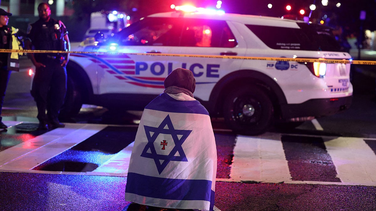 A man, with an Israeli flag with a cross in the center, kneels next to emergency personnel working at crime scene