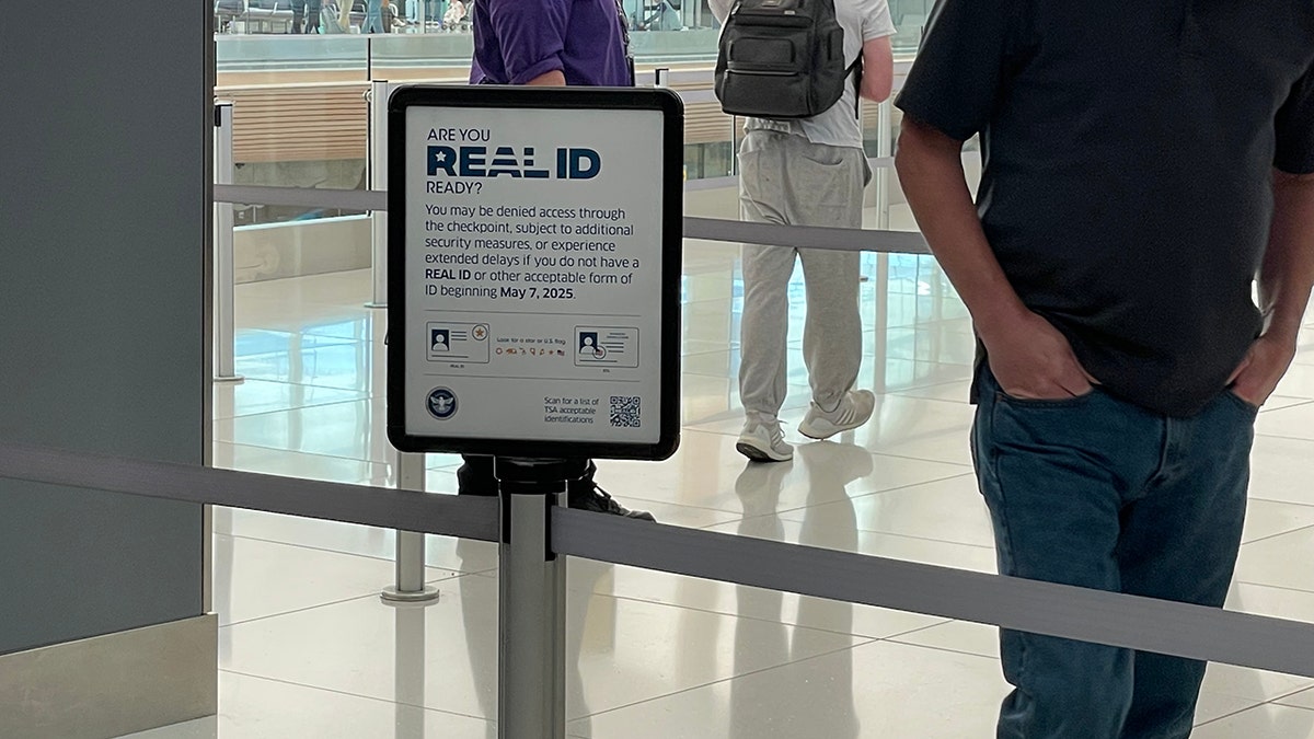 A REALID sign at the Denver airport, close up of a sign about elbow-level with people in the background of the shot. The sign reads "Are you REALID ready? You may be denied access through the checkpoint, subject to additional security measures, or experience extended delays if you do not have a REAL ID or other acceptable form of ID beginning May 7, 2025."