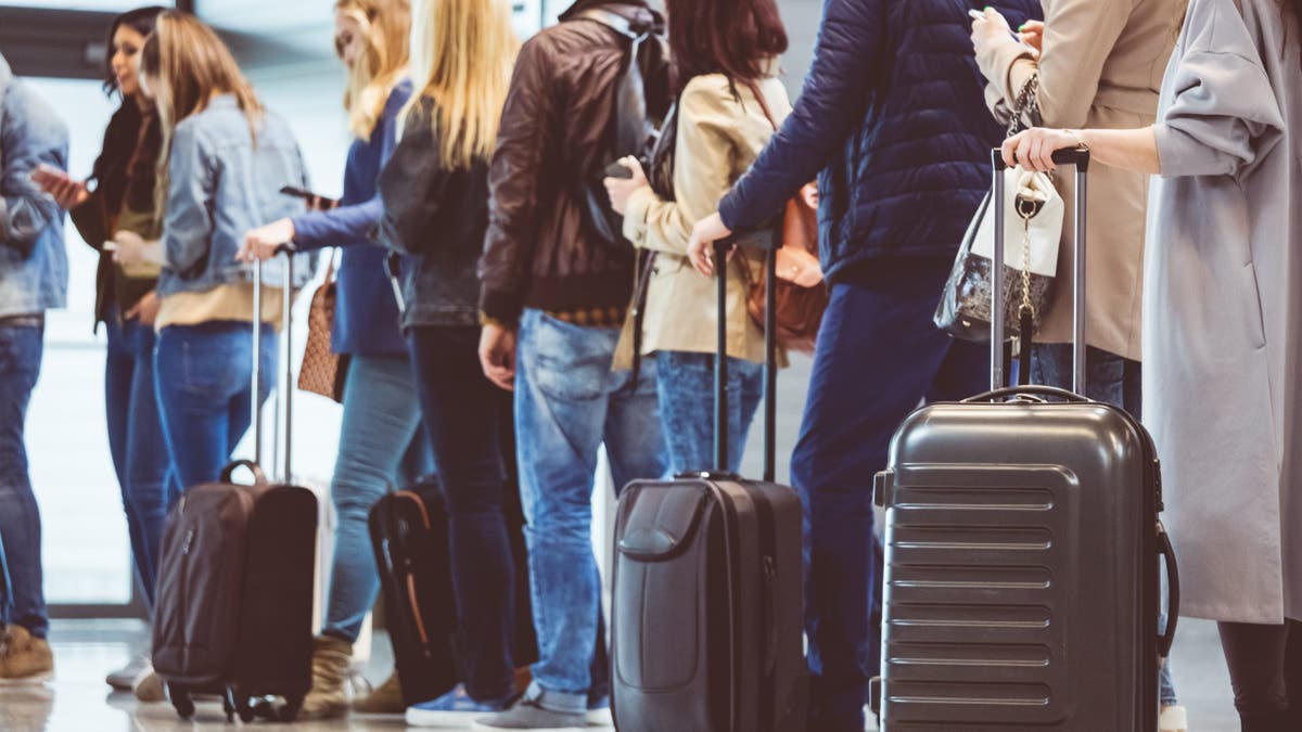 Queue of passengers pictured facing away from the camera waiting at a boarding gate at the airport. Passengers are holding suitcases and luggage as some are on their phones