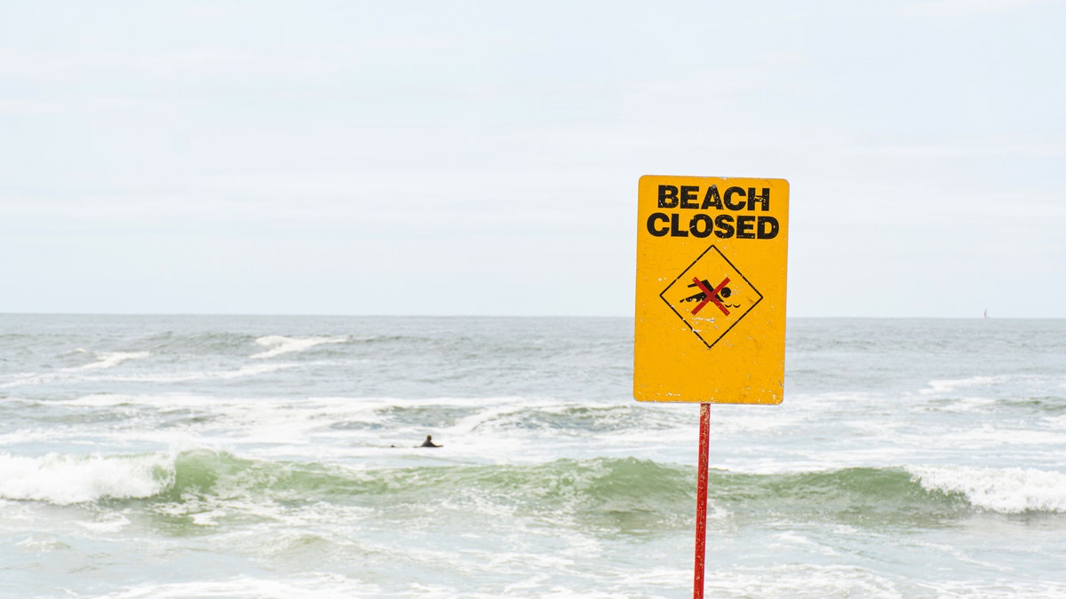 Beach closed sign standing near the ocean but someone still swimming with the board