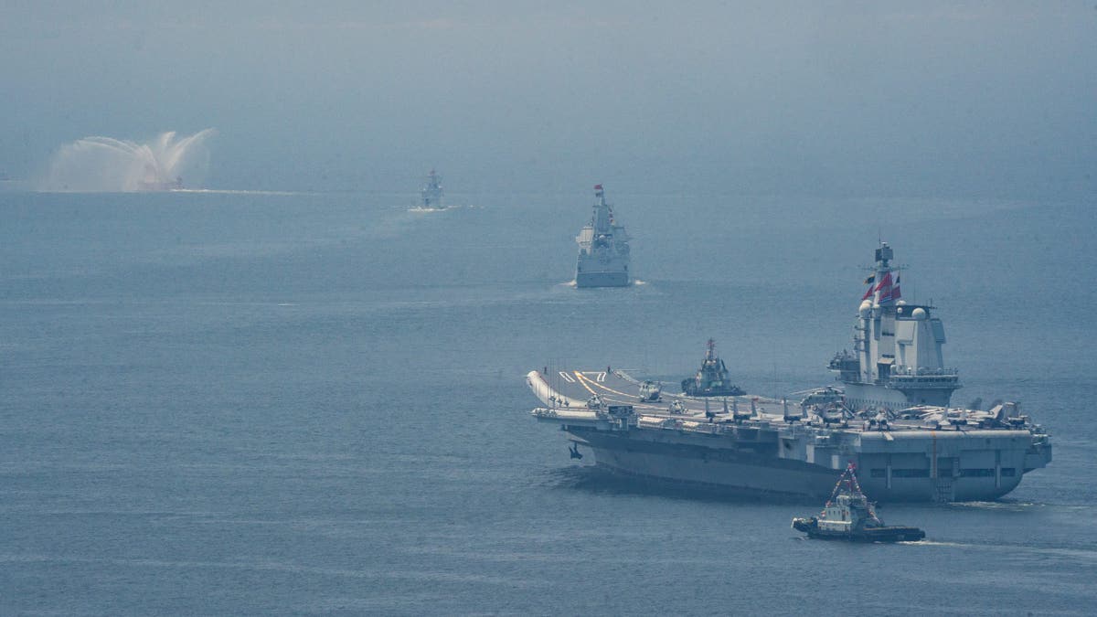 A Chinese aircraft carrier leads a formation of naval vessels during maritime operations, with support ships visible in the distance under hazy skies.