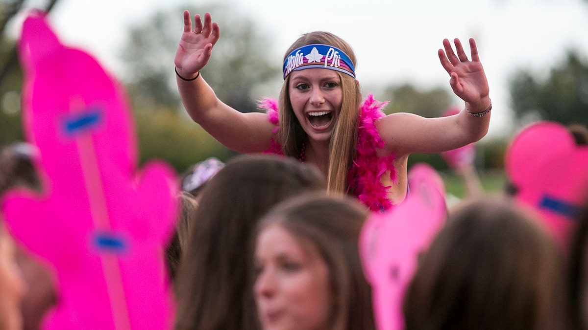 girl seen on shoulders of fellow sorority potential new members, seen with pink feather boa, headband with Greek org name, hands up yelling
