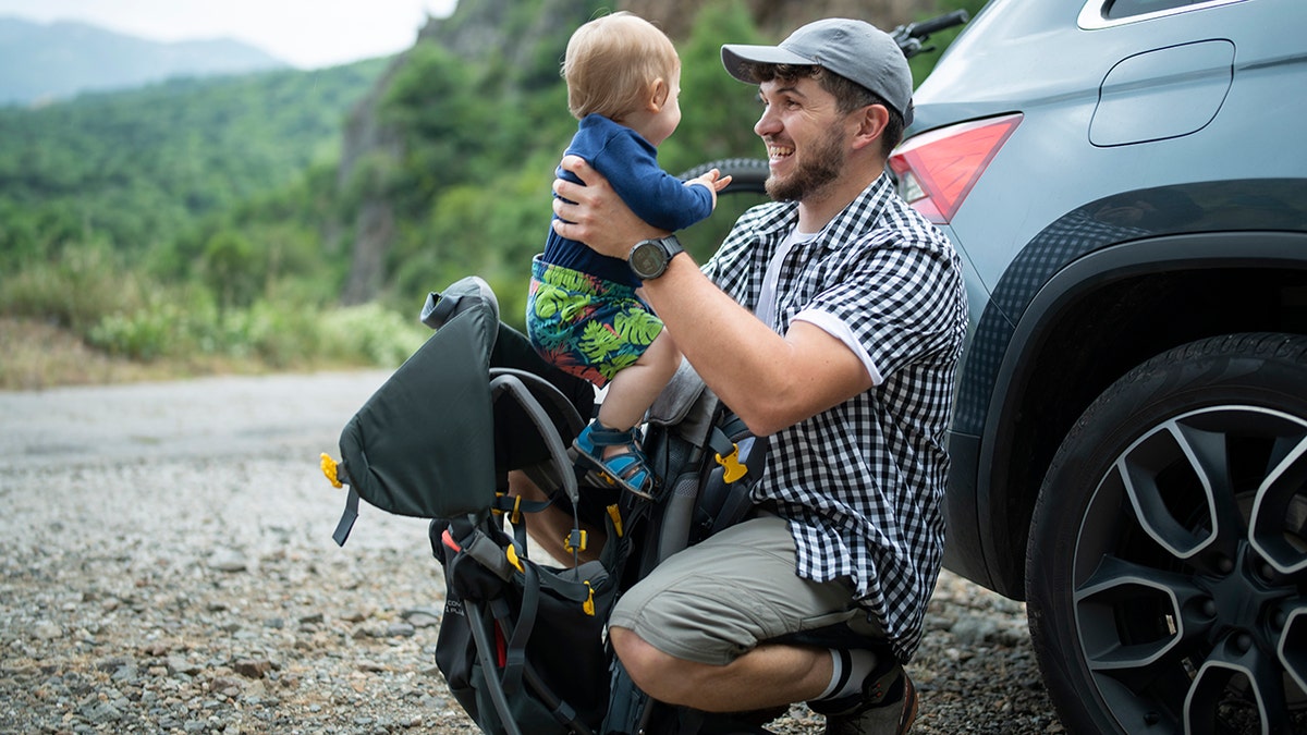 Father adjusting baby backpack carrier for going on a hike, seen holding baby up and smiling with mountains, car and bike in distance