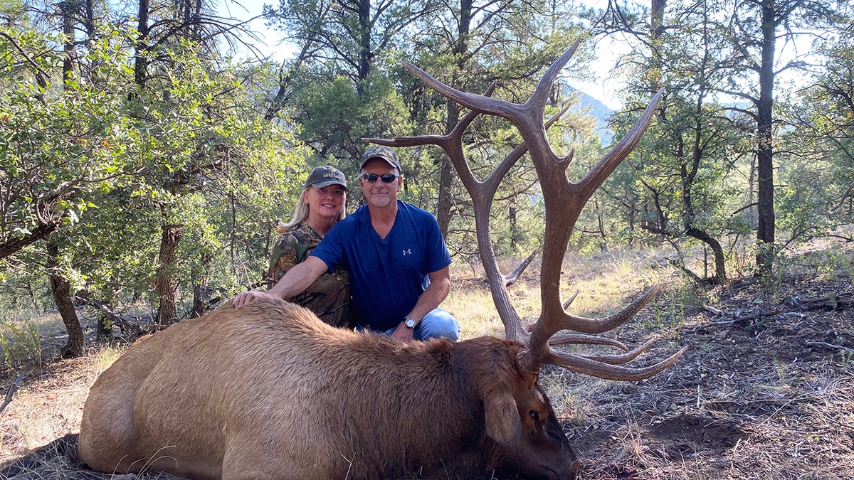 Larry Rudolph wearing a blue shirt posing in front of an elk he hunted next to Lori Milliron smiling.