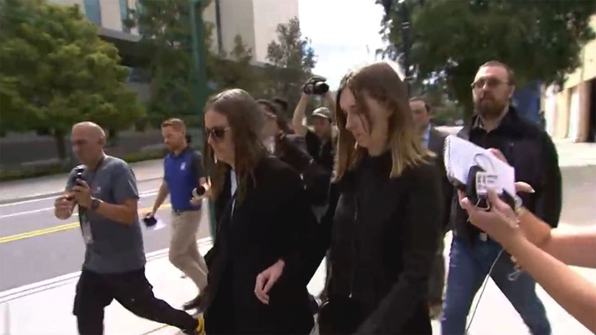 Two people in dark clothing walk arm in arm past reporters and photographers outside a courthouse.