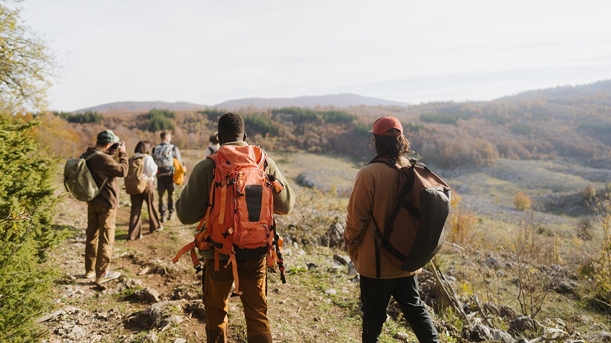 Group of hikers walking along a mountain trail overlooking a wide valley filled with autumn trees.