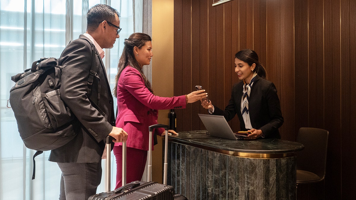 Business travelers checking in at a airport lounge reception desk, handing a phone to a smiling front desk agent.