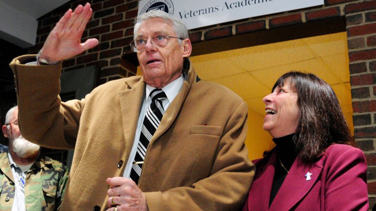 Robert Fuller Jr. in tan coat and tie waves at crowd inside veterans facility