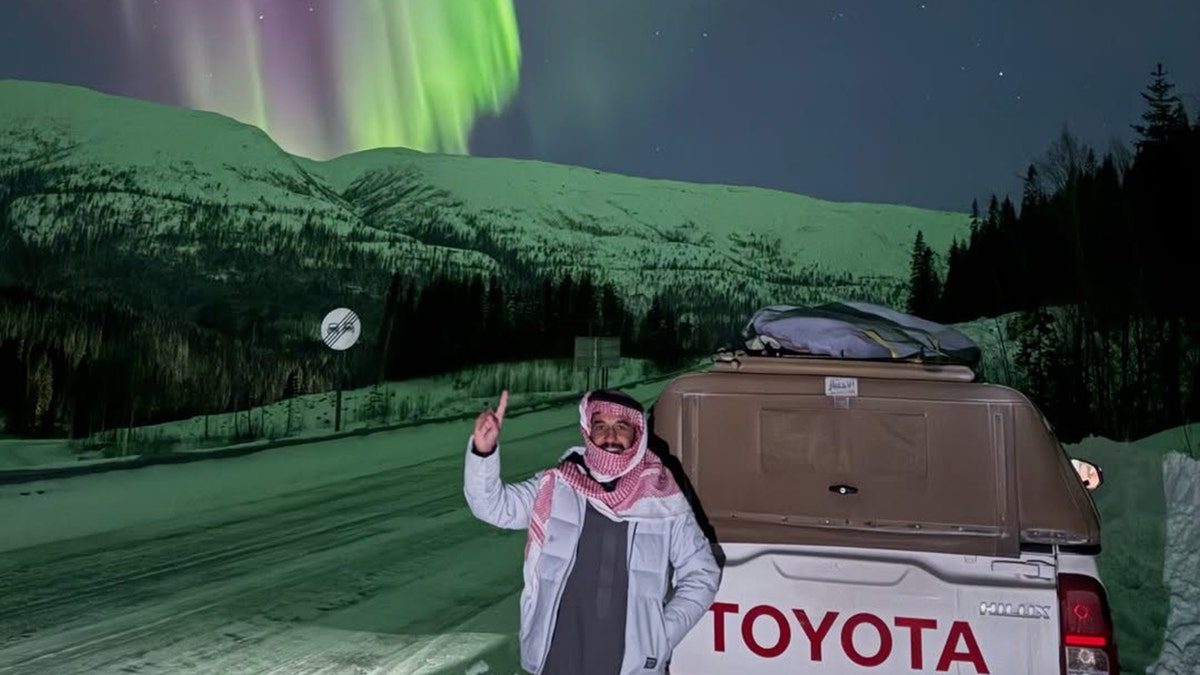 Malik Al-sultan standing beside a Toyota pickup truck under the northern lights on a snowy mountain road at night.