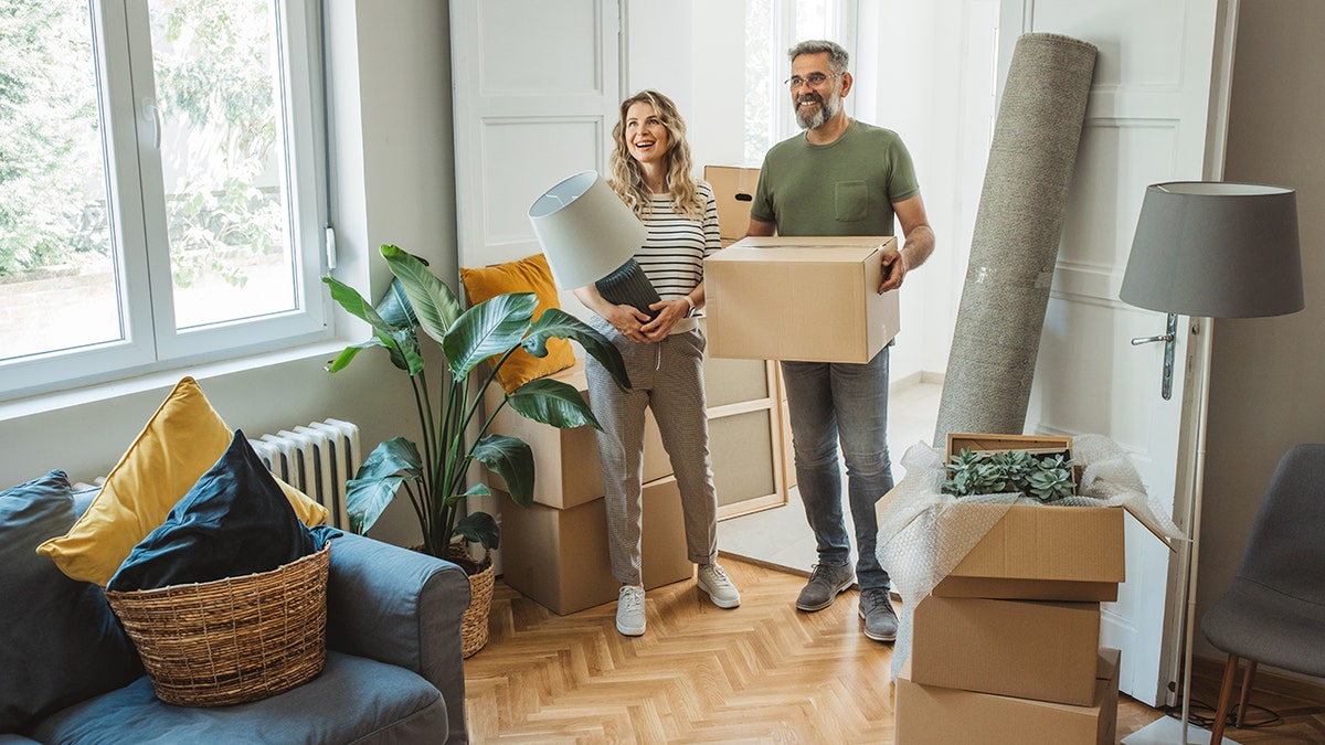 Middle-aged couple standing in a bright living room surrounded by moving boxes while unpacking items in their new home.