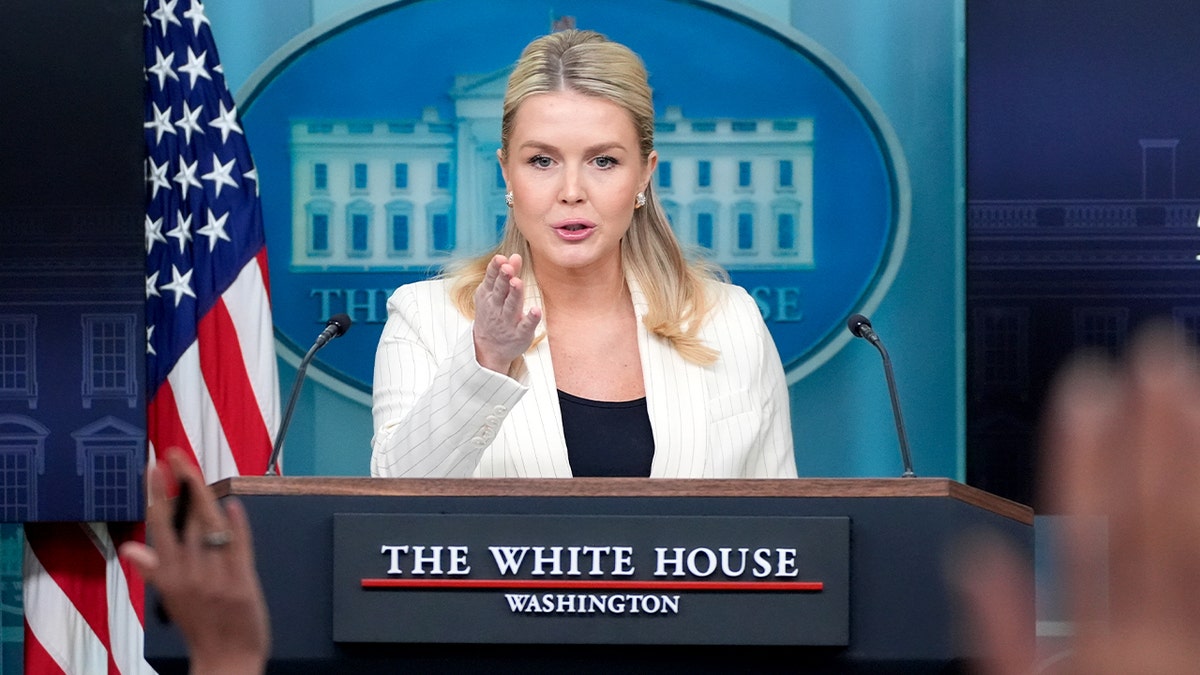 Karoline Leavitt stands at a lectern addressing journalists inside a government briefing room.