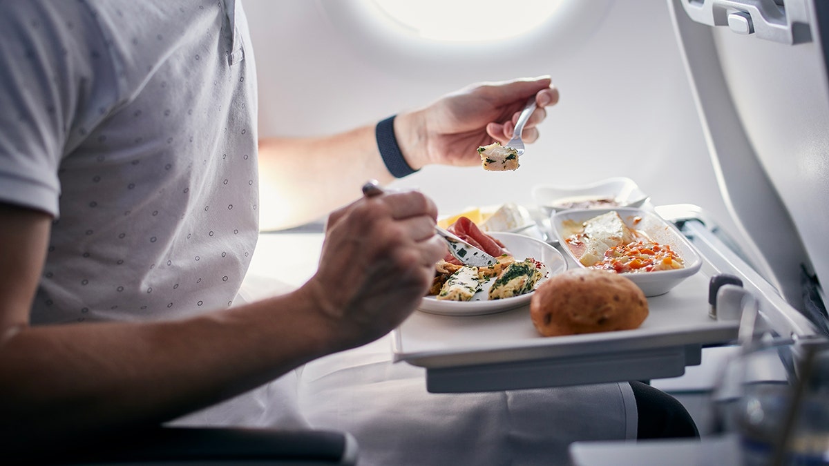Passenger eating an in-flight meal on an airplane, with a tray table holding plated food and bread roll.