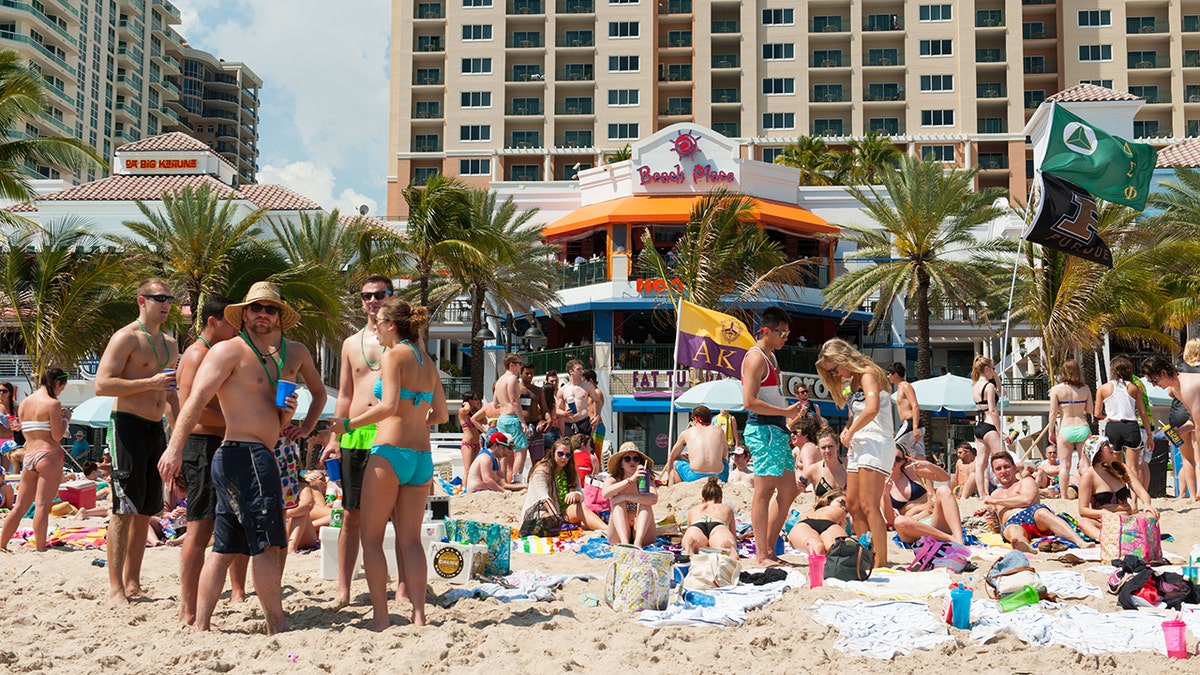 Crowded Fort Lauderdale beach in front of Beach Place with college students sunbathing, socializing, and palm trees with high-rise beachfront hotels in the background.
