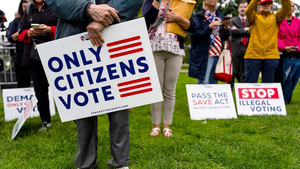 People gathered outdoors hold signs and watch a speaker address a crowd near the U.S. Capitol.