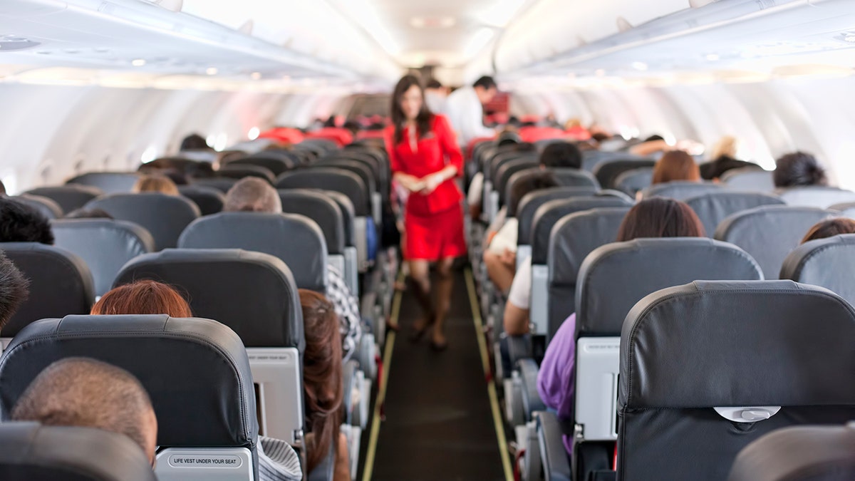 A blurred flight attendant wearing red is seen standing in the cabin of a commercial flight.