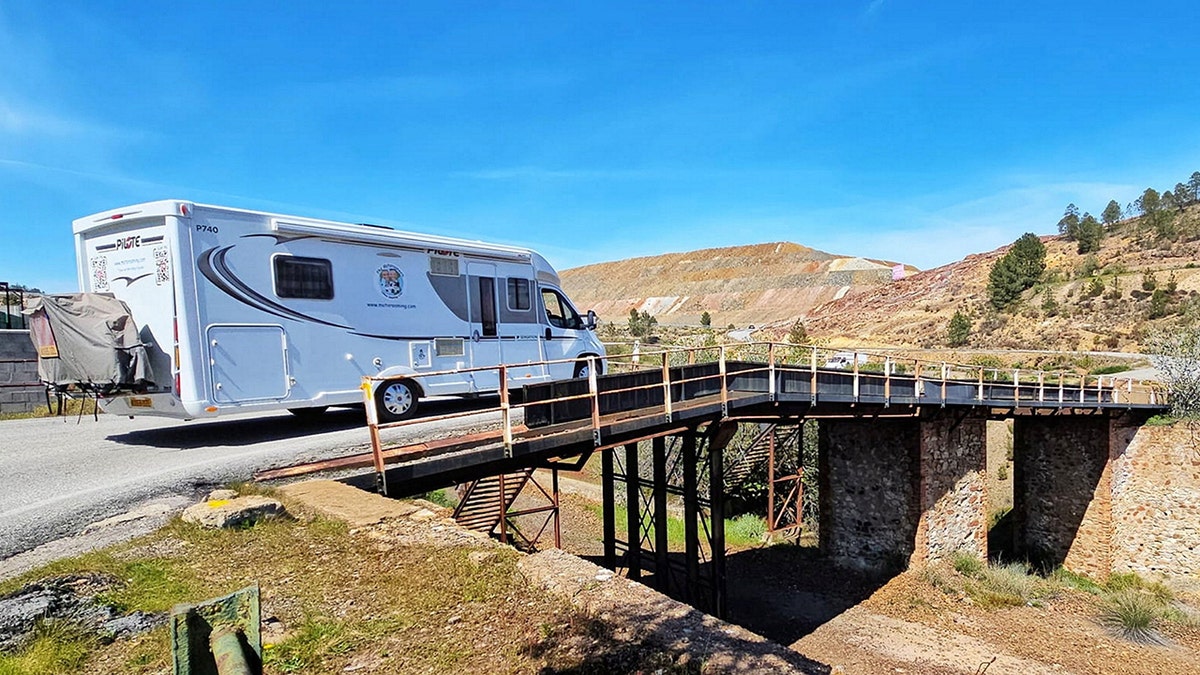 Motorhome crossing a narrow metal bridge in a rugged desert mining landscape under a clear blue sky.