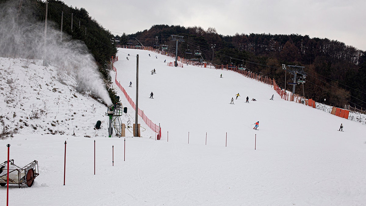 Skiers and snowboarders descend a wide snow-covered slope at a ski resort, with a snowmaking machine blowing artificial snow along the side of the run.
