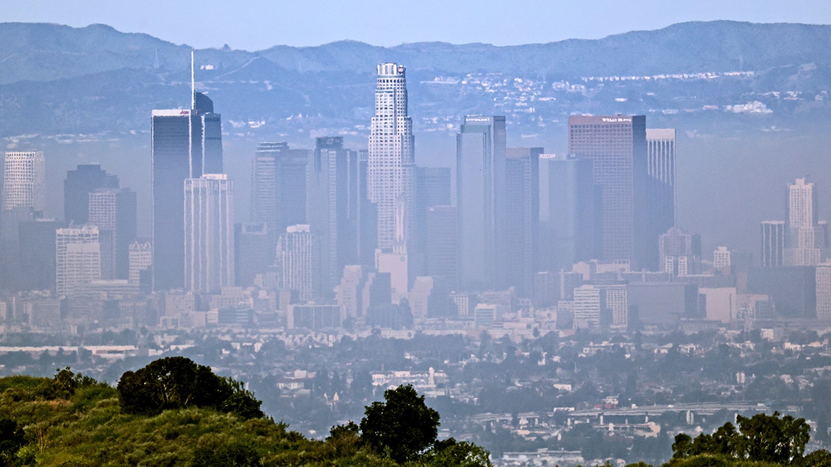 Los Angeles skyline with downtown skyscrapers partially obscured by heavy smog and haze, viewed from a distance with hills in the background.