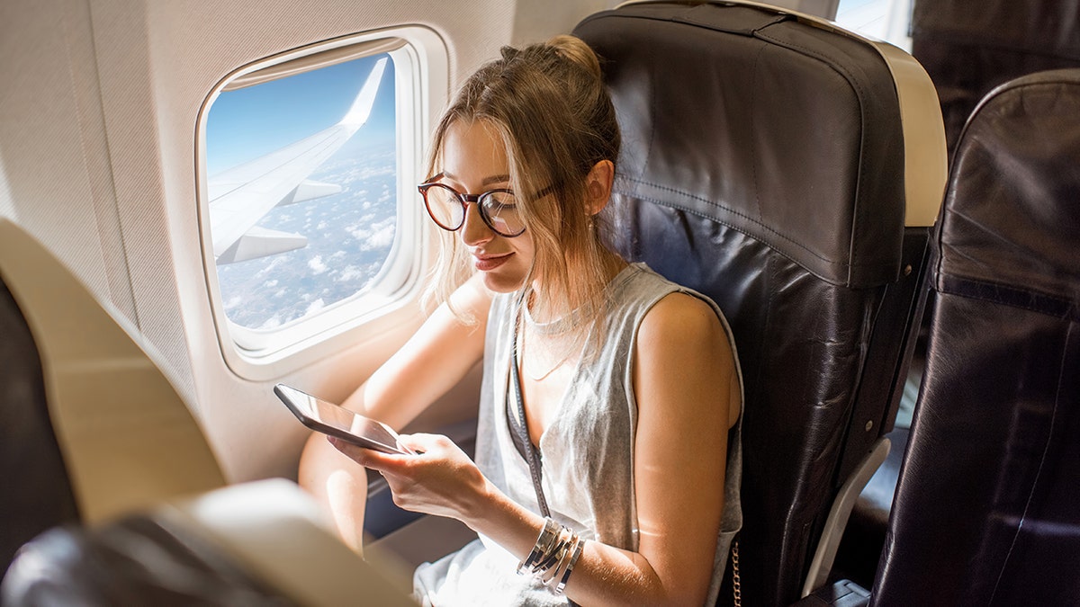A woman looking at her phone on an airplane with a view of the sky through the window.