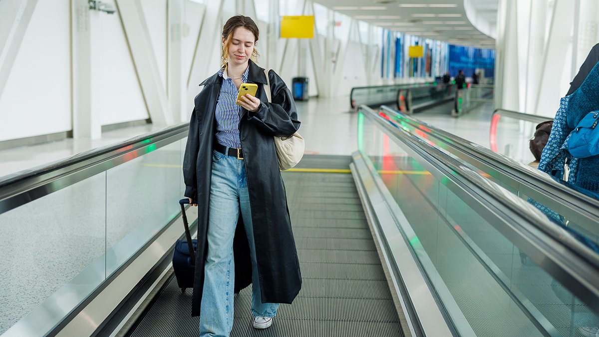 Woman walking on an airport moving walkway while checking her smartphone and pulling a rolling suitcase.