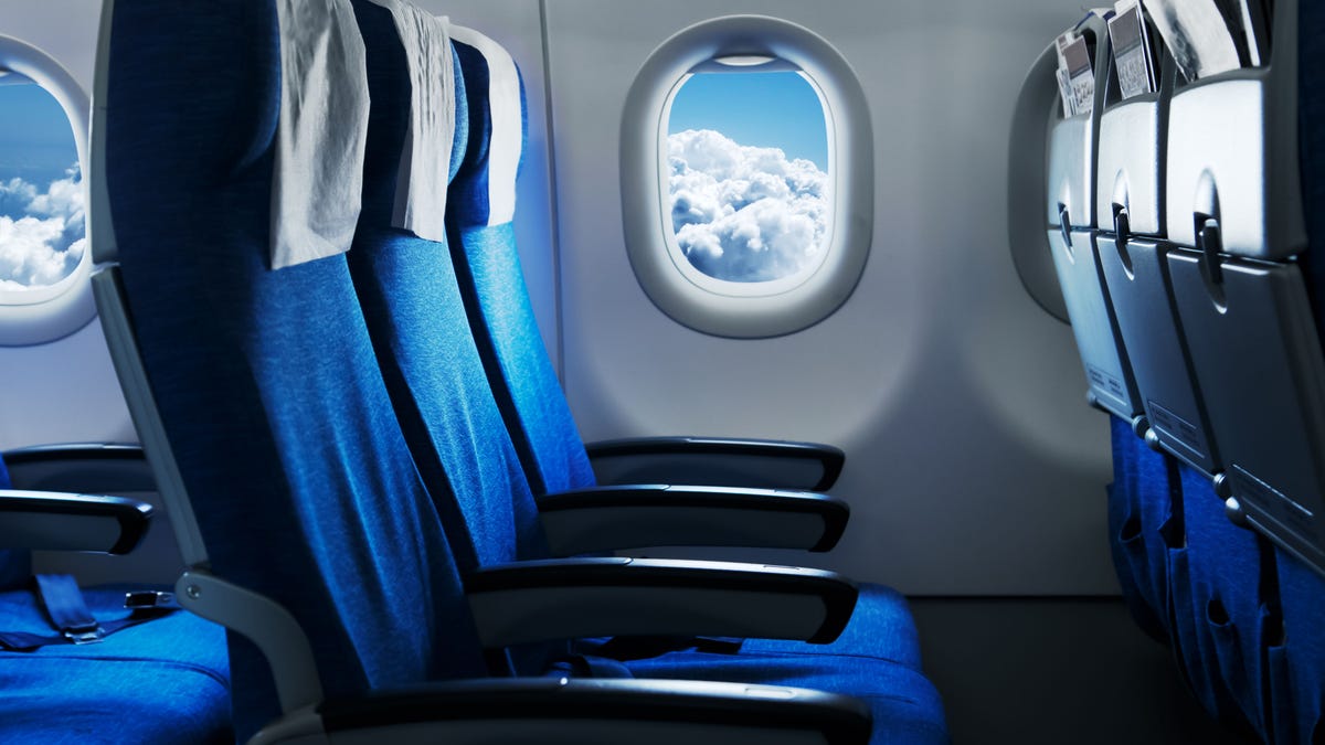 Empty airplane seats with blue sky and clouds visible through window.