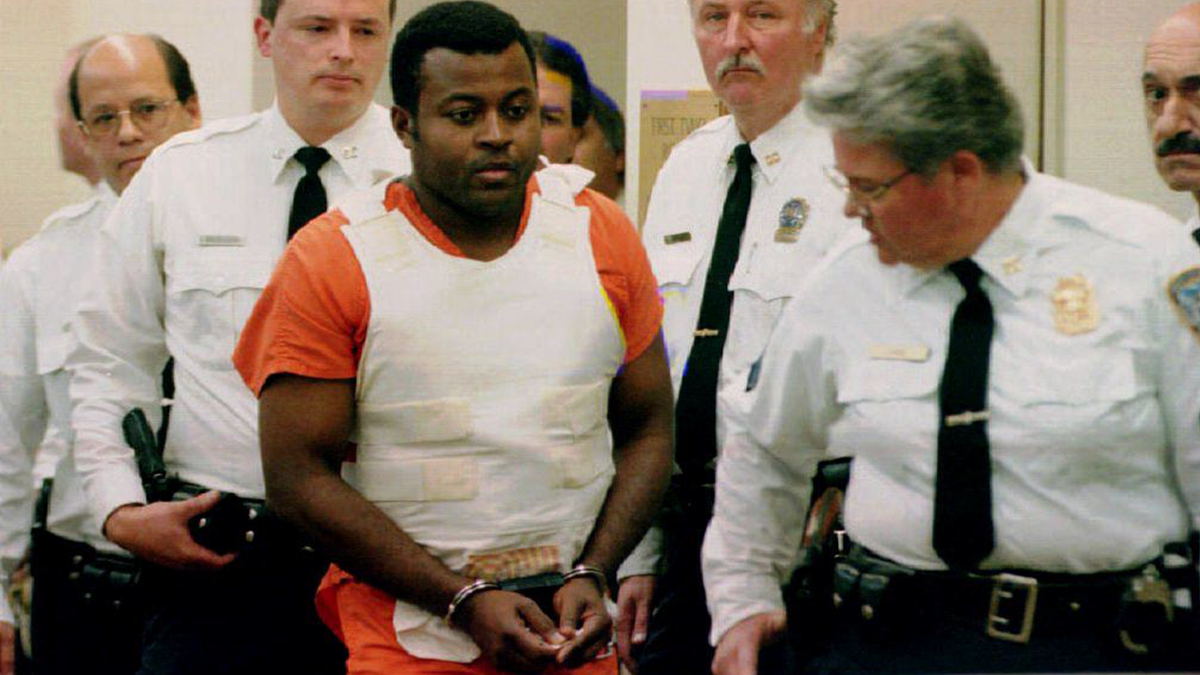 Colin Ferguson in handcuffs, an orange jail jumpsuit and white bulletproof vest, surrounded by court officers