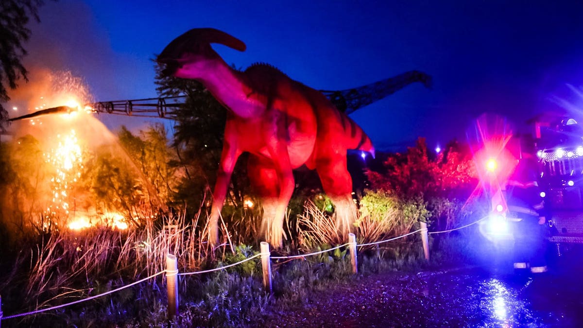 Dinosaur replica in Field Station at night as firefighters spray water on a nearby blaze, with emergency lights flashing.