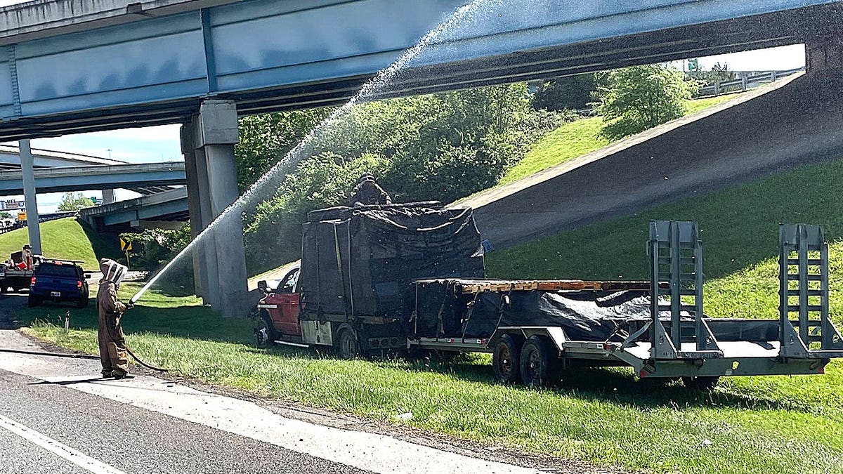 A person in a tan bee suit sprays a jet of water toward a dark-colored trailer parked near a highway overpass.