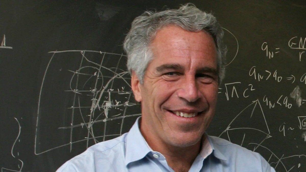 A close-up of Jeffrey Epstein smiling with a light blue collared shirt standing in front of a chalkboard.