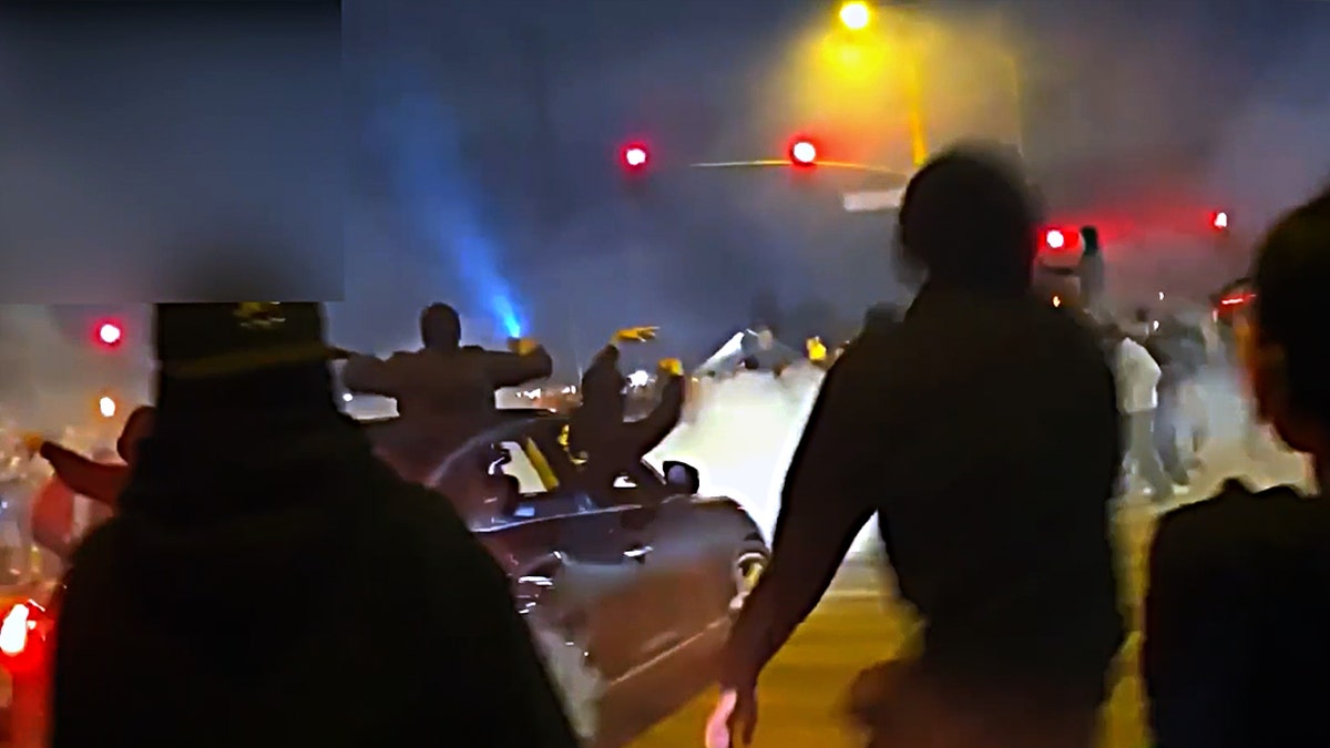 A car with people hanging out of it drifts through an intersection as people look on.