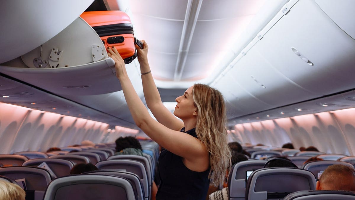 Woman placing pink hard-shell suitcase in airplane overhead storage compartment