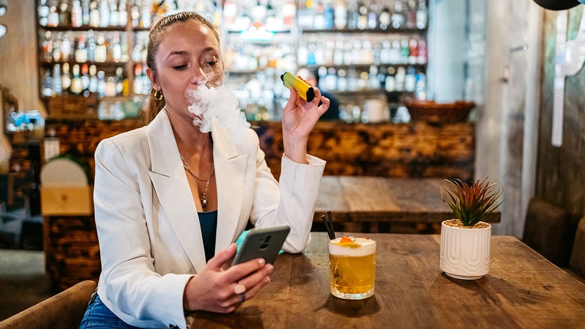 Beautiful young woman vaping and texting on her smartphone in a bar.