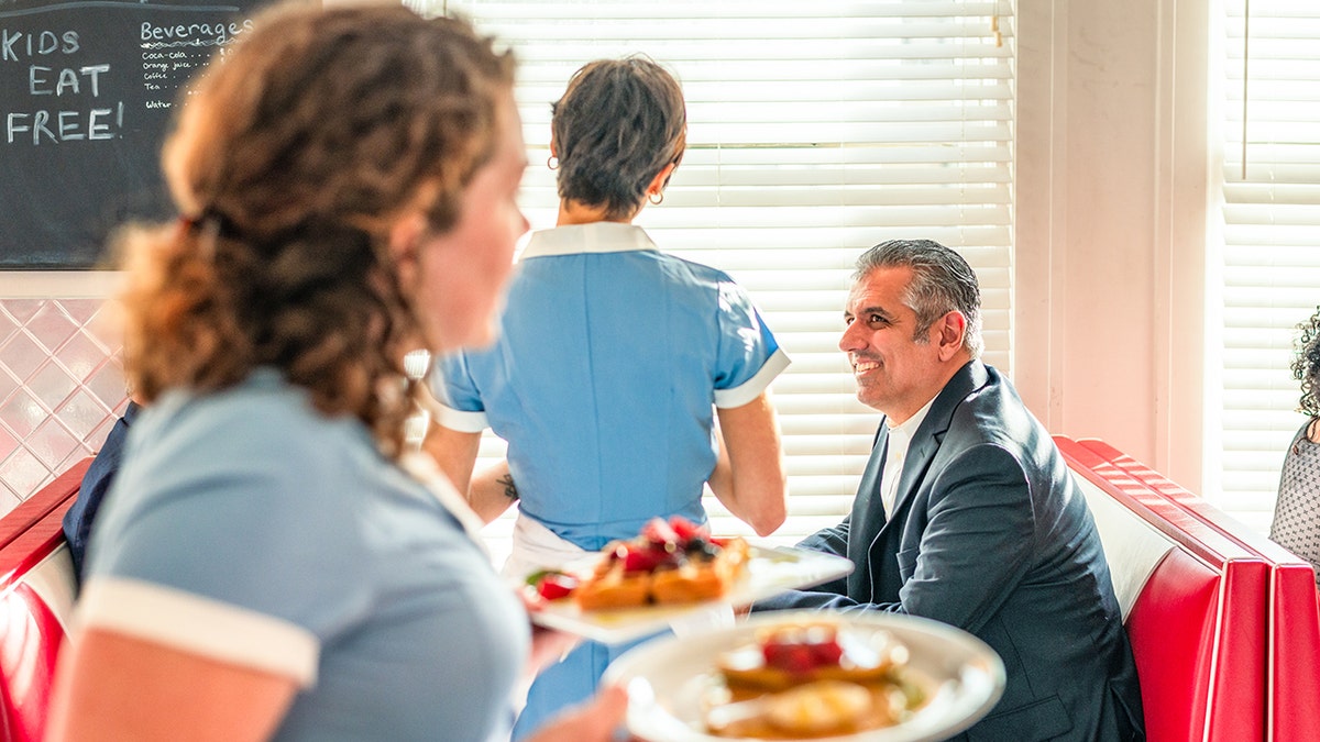 Two waitresses serving breakfast in a busy diner with a businessman seated at a booth