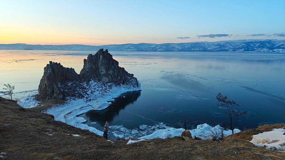 A rocky island in a partly frozen lake with snow along the shore and a sunset sky.