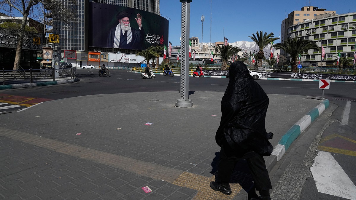 A woman walking across a nearly empty public square with a large billboard of Ayatollah Ali Khamenei in the background