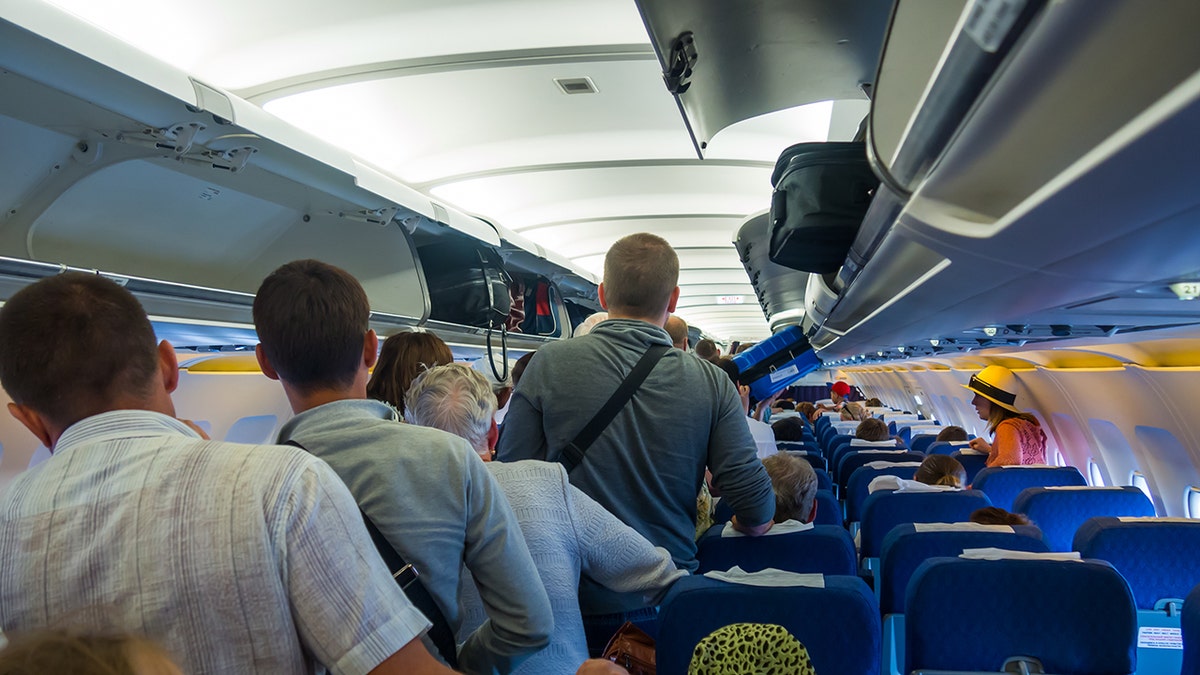 Passengers standing in a crowded airplane aisle storing carry-on bags in overhead compartments