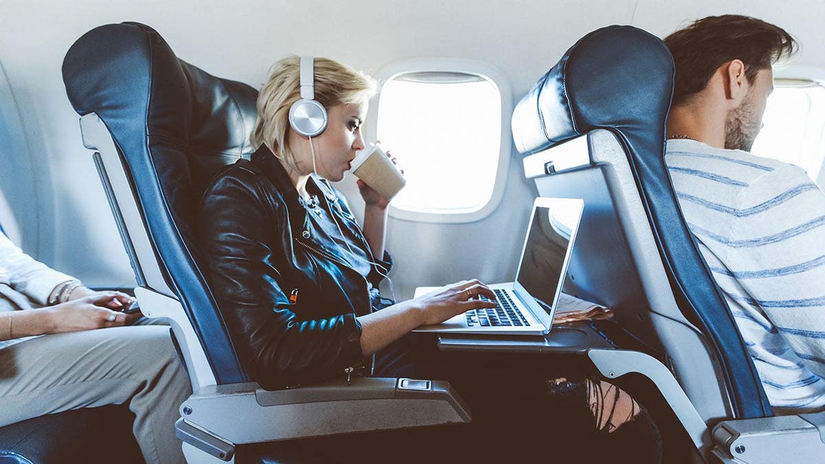 Young blonde woman sitting inside an airplane using a laptop and drinking coffee