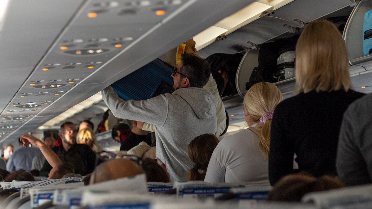 Passengers stowing carry-on luggage in overhead bins inside a crowded airplane cabin