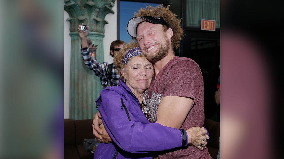 Daredevil Andy Lewis is wearing a black visor and maroon T-shirt as he has curly, light brown hair and a short beard in this photo, where he hugs his mother Lynn after setting a world record in Las Vegas