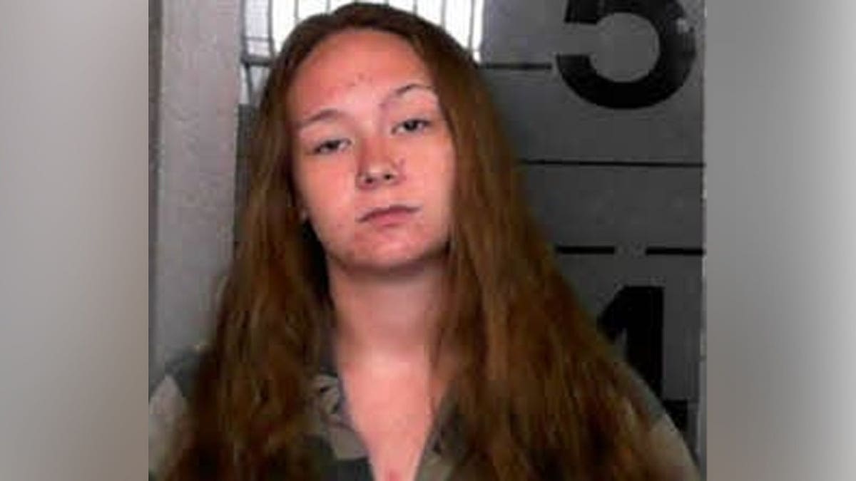 Booking photo of a young woman with long reddish-brown hair standing against a height chart background.