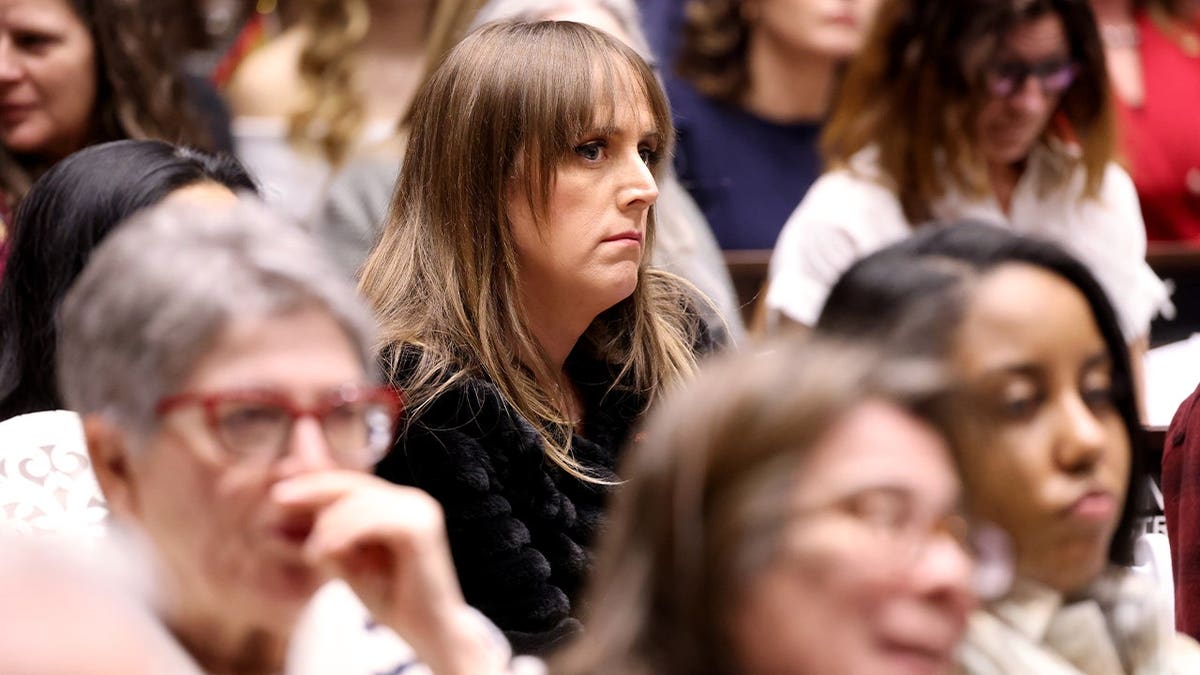 Woman with long straight hair and bangs, wearing a black textured jacket, sits among a crowd of other attendees indoors.