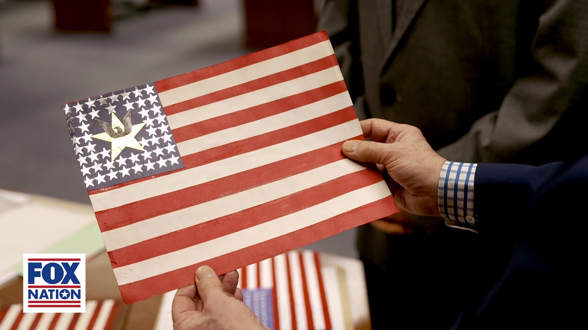 A hand holds a proposed 50-star U.S. flag design with a white eagle displayed prominently among the stars in the blue canton.