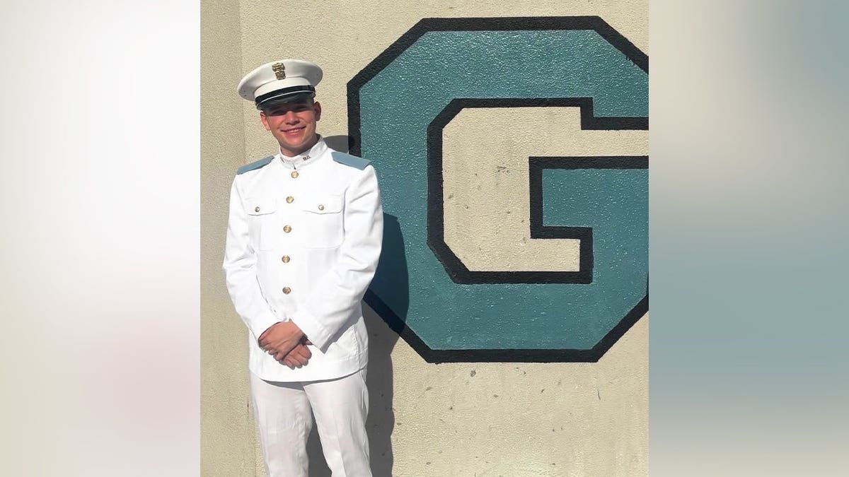 A young man in a white Citadel cadet dress uniform and cap stands smiling beside a large painted