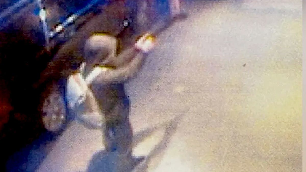 A hooded man wearing a backpack and aiming a gun with a silencer on a new york city sidewalk