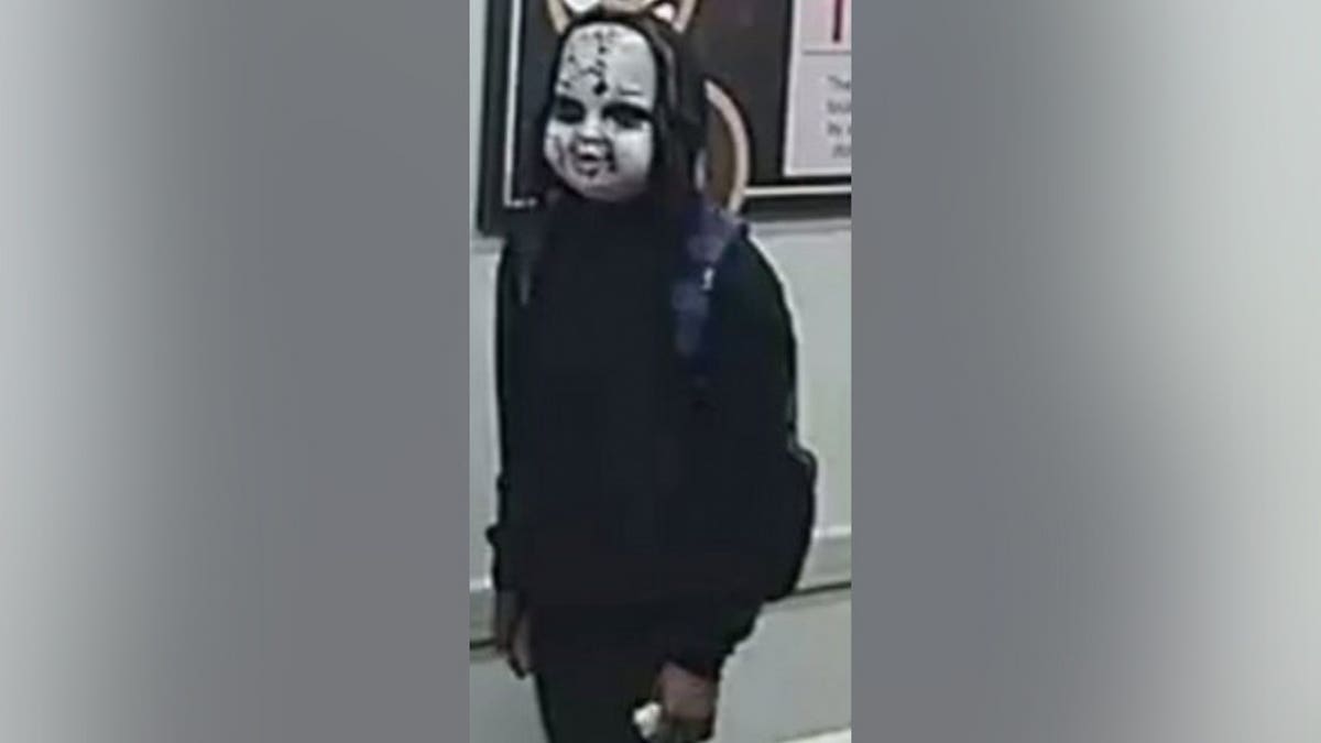 Surveillance still of a person in a black hoodie and pants wearing a white cracked doll mask, standing indoors.