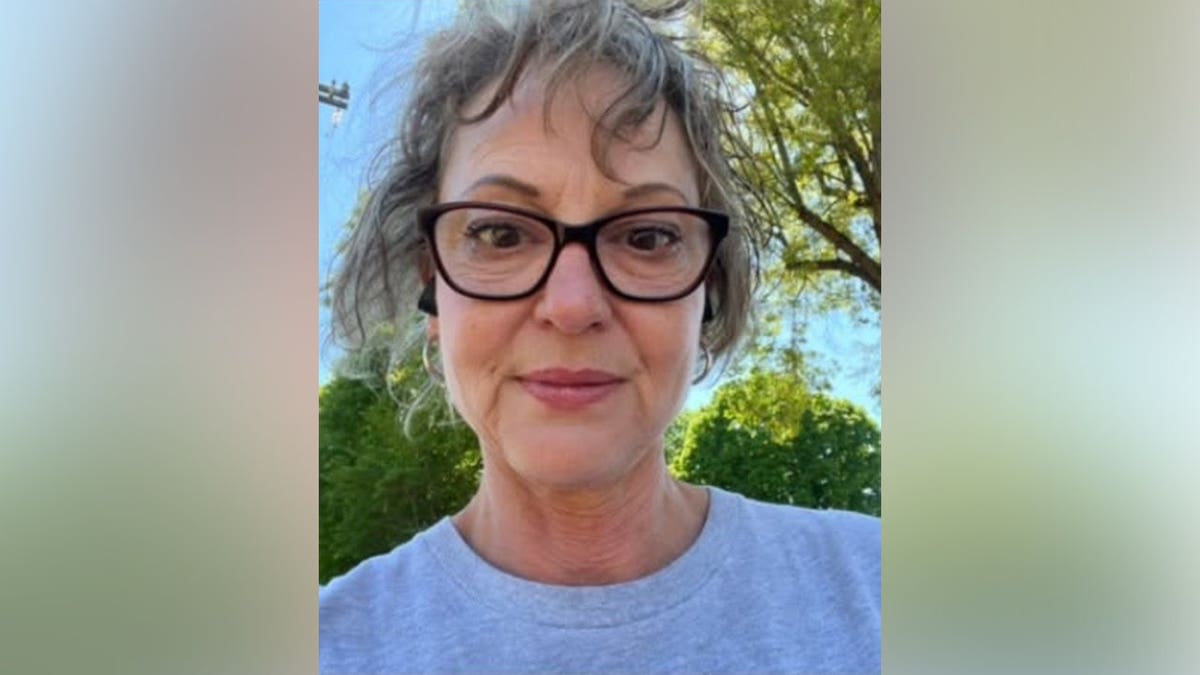A woman with gray curly hair and dark-framed glasses wearing a gray T-shirt, smiling slightly outdoors with green trees behind her.