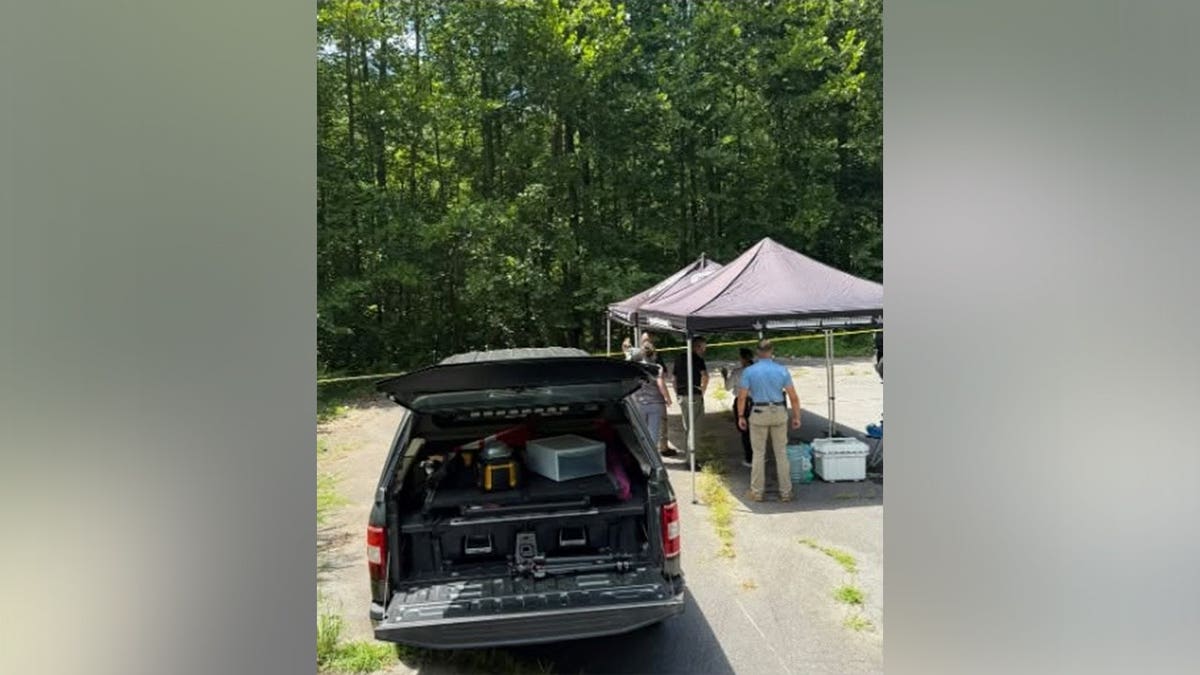 Law enforcement officers stand under a gray canopy tent beside an SUV with its rear hatch open, gathered along a paved area at the edge of dense woods marked with police tape.