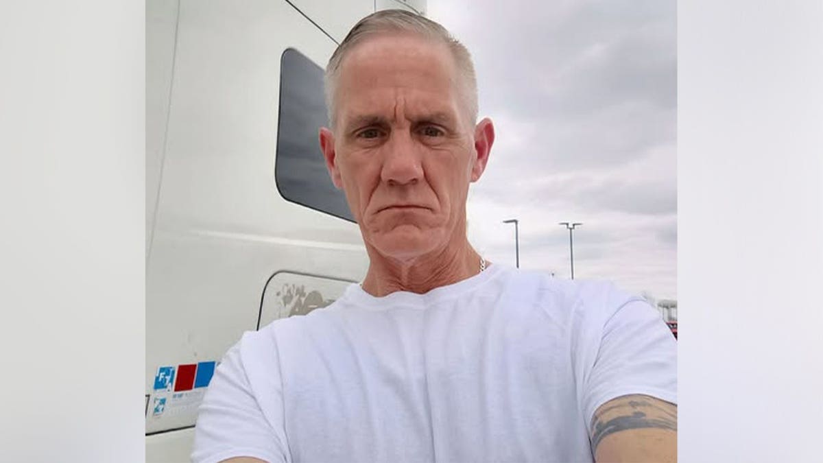 Fredrick Lytton, a 56-year-old man with short gray hair, wears a white T-shirt while standing in front of a semi-truck cab.