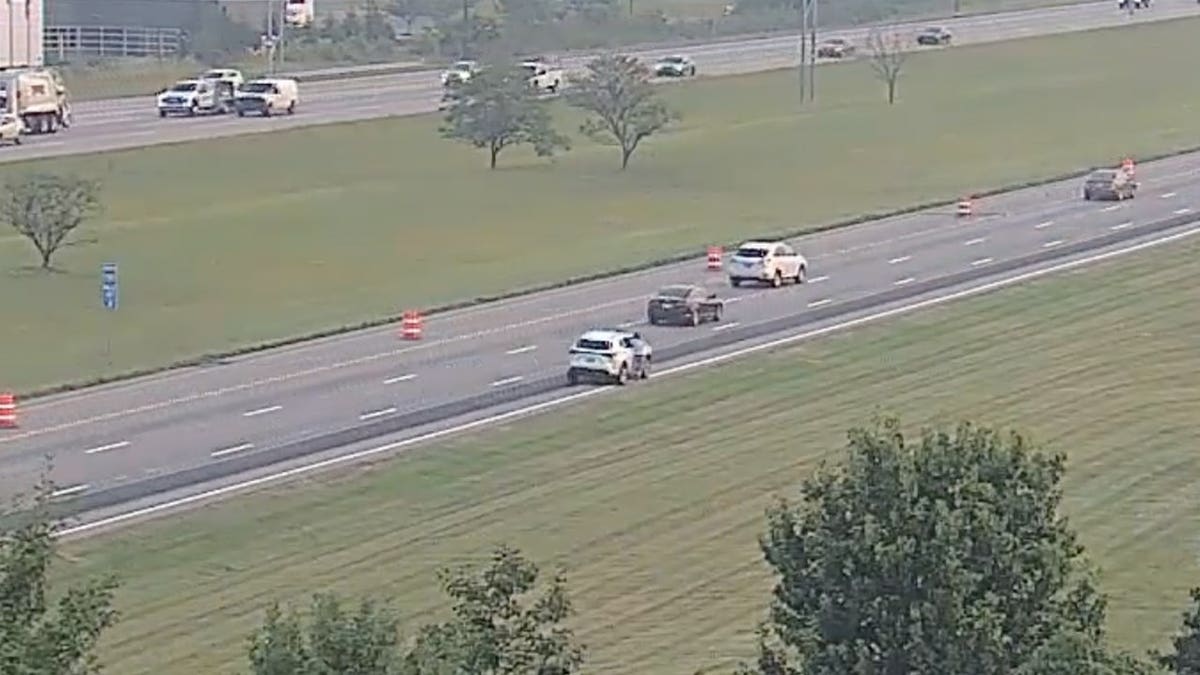 Highway camera view of vehicles traveling on a multi-lane interstate bordered by grass and trees.